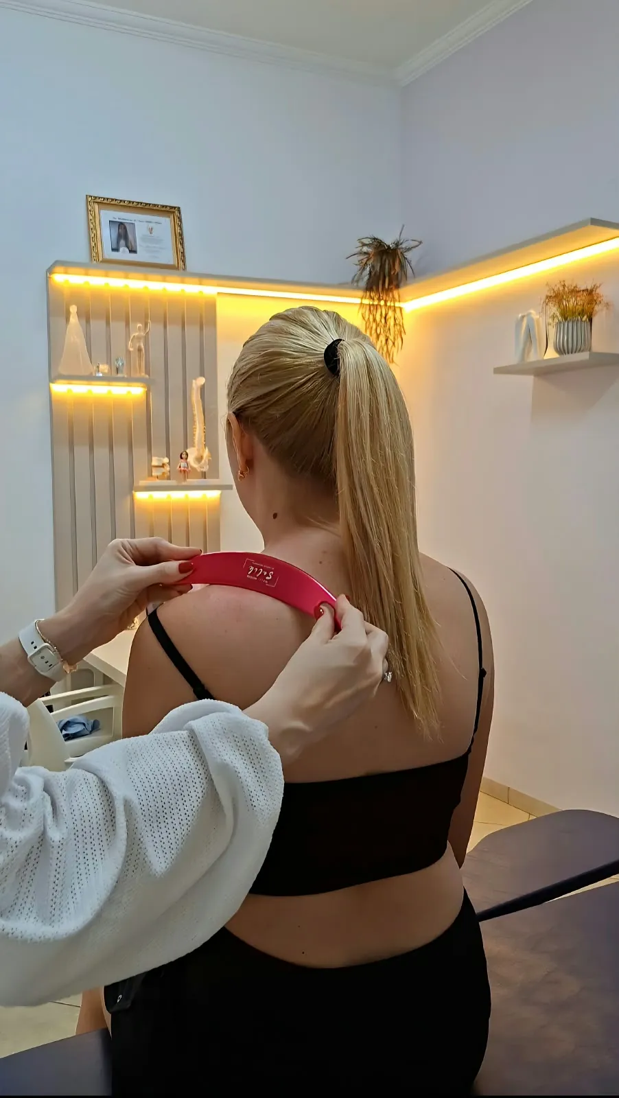 Dra. Gisela aplicando um instrumento de liberação miofascial no ombro de uma paciente, no consultório com iluminação quente