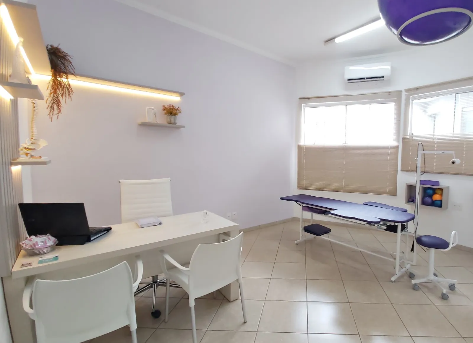 Sala de atendimento clara, com mesa, cadeiras brancas, maca roxa e janela